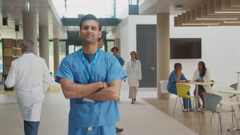 portrait of male doctor wearing scrubs folding arms and standing in busy hospital