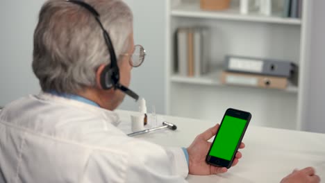 el médico masculino consulta al paciente utilizando un teléfono móvil con pantalla verde, llave de croma y auricular. diagnóstico de examen a distancia remoto en línea por videoconferencia, chat de teleconferencia. telemedicina.