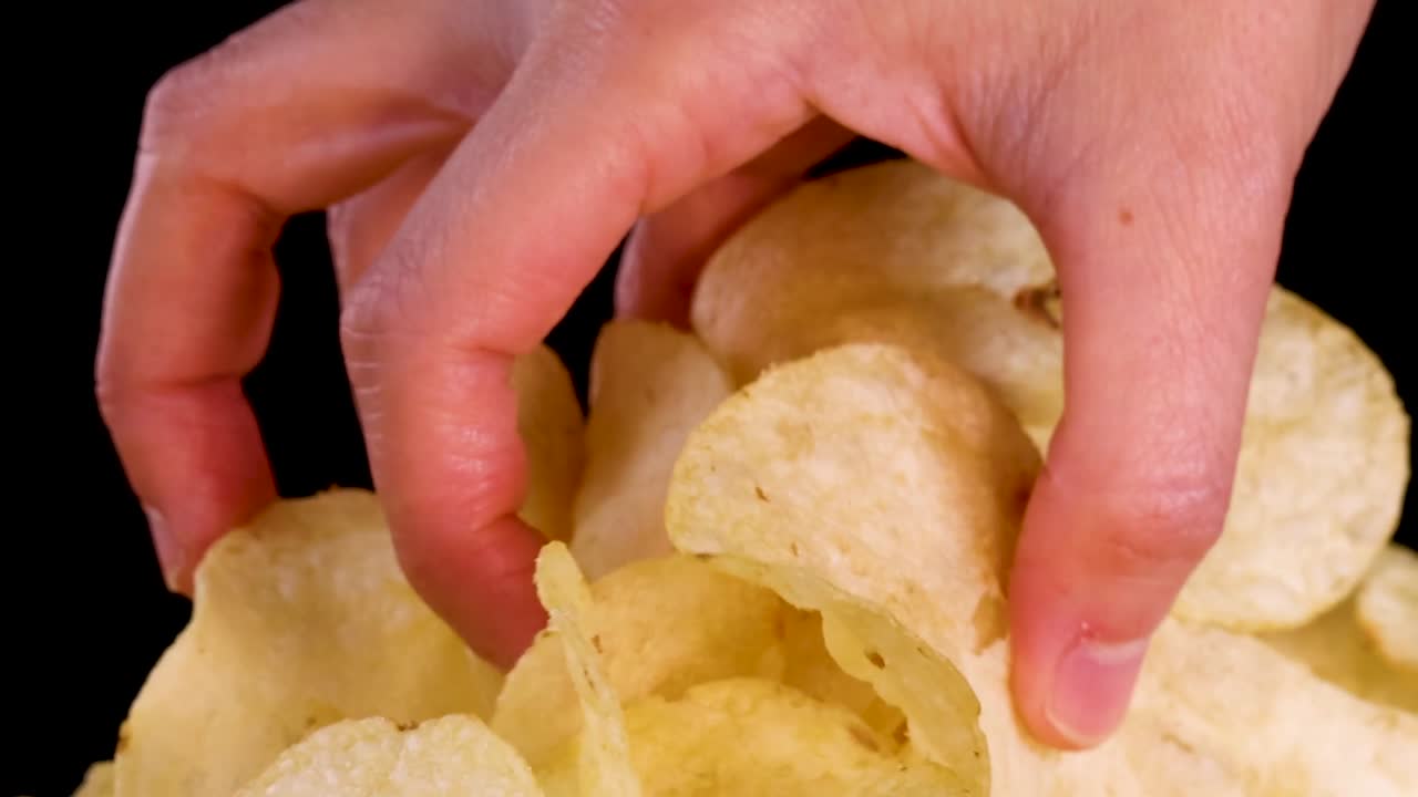 A human hand reaches into a pile of golden potato chips against black background