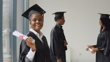 one young african-american woman stands in the foreground and looks at the camera with a diploma in her hands. young african american man and woman communicate with each other in the background.