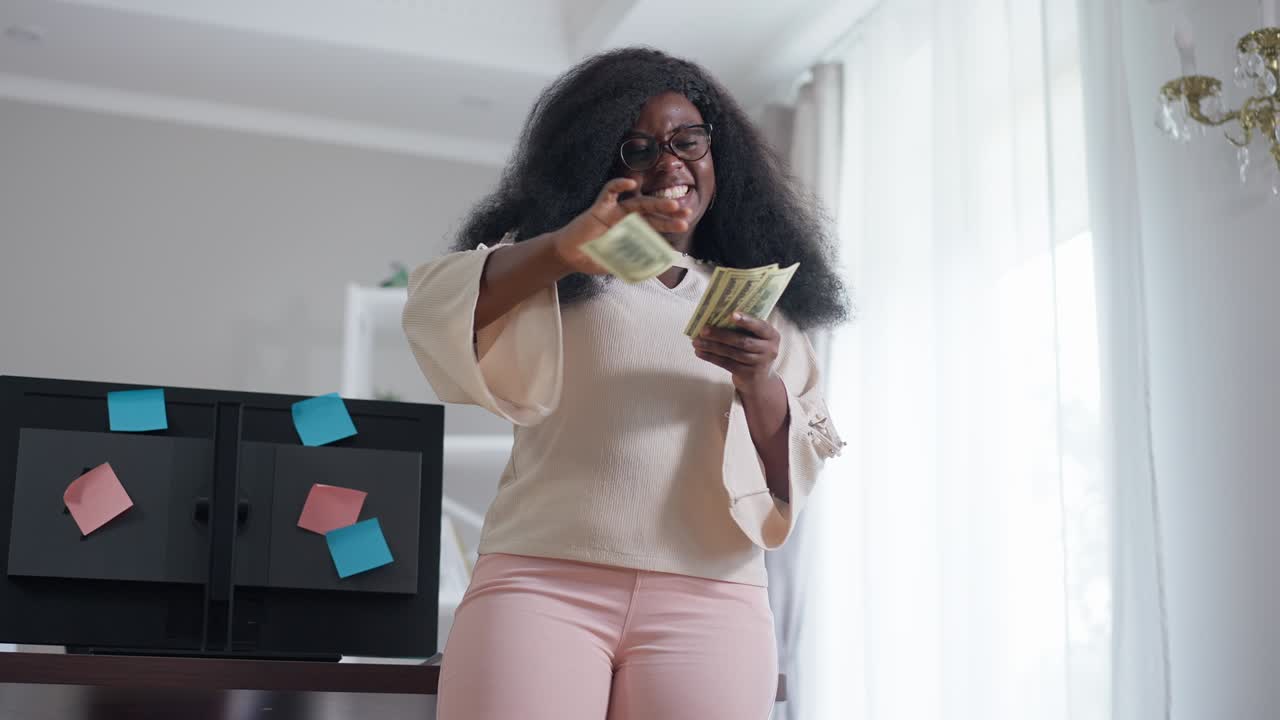 Happy young African American rich woman scattering money indoors in slow motion smiling. Portrait of wealthy successful freelancer rejoicing success standing in home office. Richness and wealth.