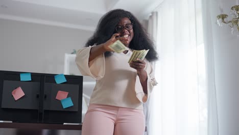 happy young african american rich woman scattering money indoors in slow motion smiling. portrait of wealthy successful freelancer rejoicing success standing in home office. richness and wealth.