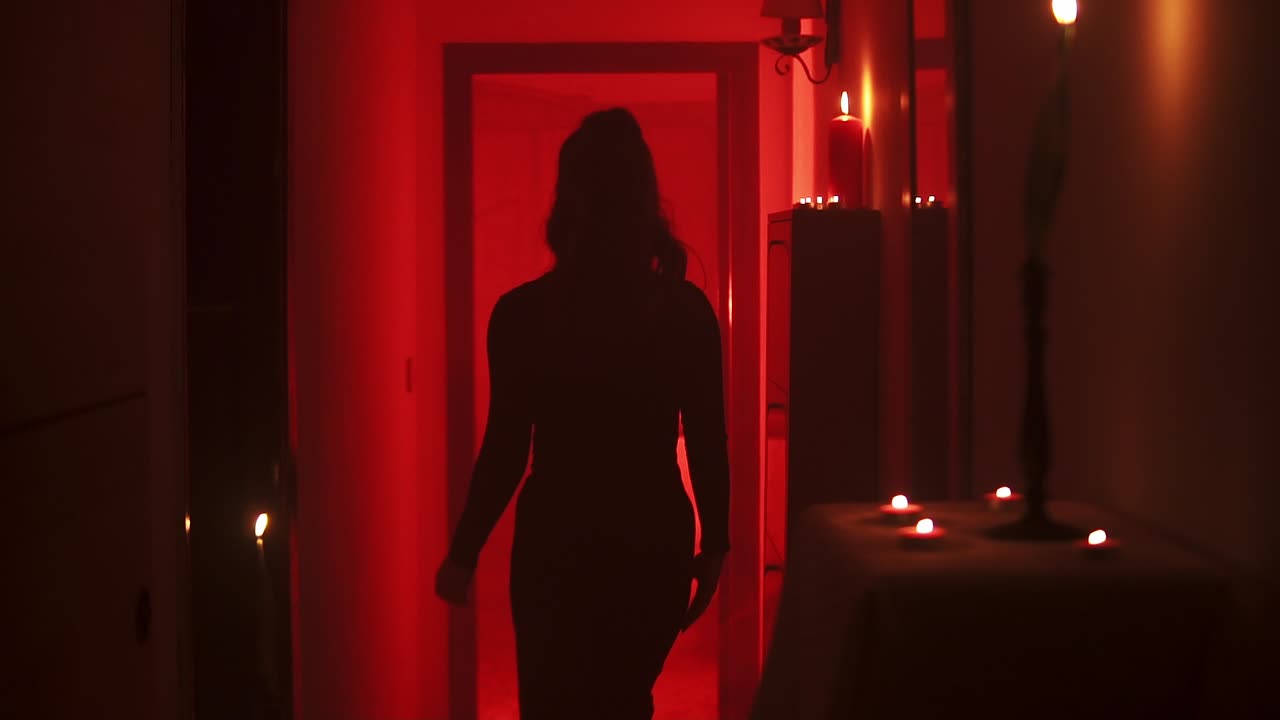Cinematic slow motion shot of a woman walking seductively into a room on a stage with red mood lighting and burning candles