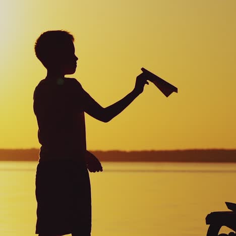 ein kleines kind hält ein origami aus papierflugzeug in der hand und spielt bei sonnenuntergang auf dem hintergrund eines sees. ein junge spielt leidenschaftlich mit seinem papierspielzeug
