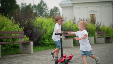 a younger and older brother joyfully run around a scooter outdoors, both holding onto the handlebar as they play together