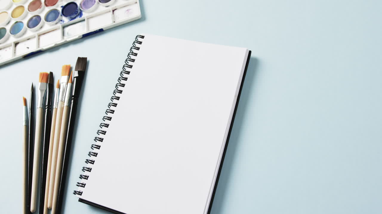 primer plano del cuaderno, pinceles y pinturas sobre fondo azul, en cámara lenta