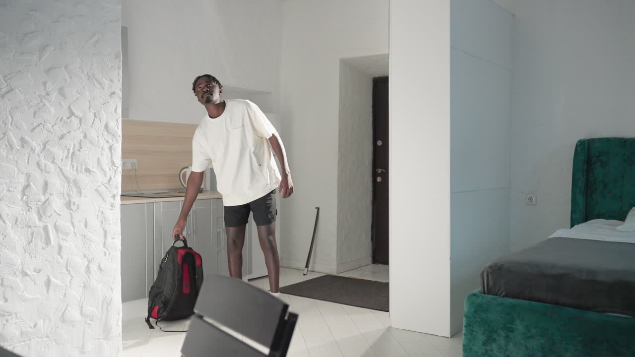 Man in casual wear walking into modern apartment with gray kitchen, refrigerator, and green velvet bed in the background. White walls and light interior create a spacious, relaxed atmosphere