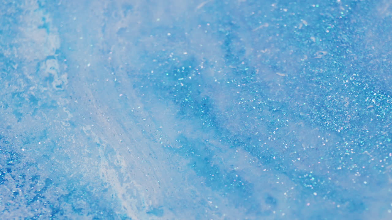 Macro shot of blue glitter ink being pushed aside by a milky liquid introduced from the top right, creating soft fluid movement