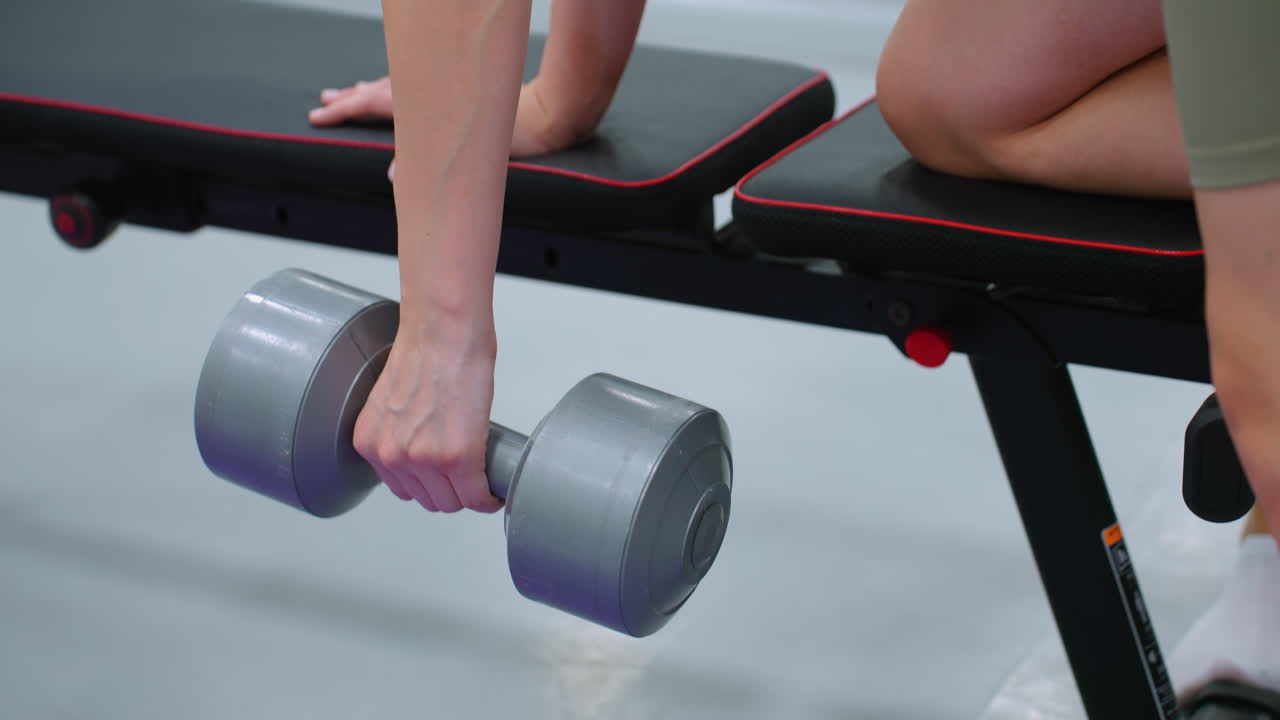 운동 벤치에 손과 무을 고 덤벨을 들어 올리는 여성의 클로즈업, 근육 참여, 손잡이 힘, 통제된 움직임에 초점을 맞추고, 다른 사람이 근처에 서 있습니다.