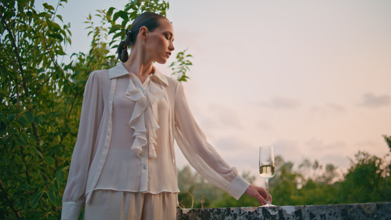 매력적인 소녀는 저녁 하늘 앞의 돌 울타리 발코니에서 와인 글래스를 찾고 있습니다.