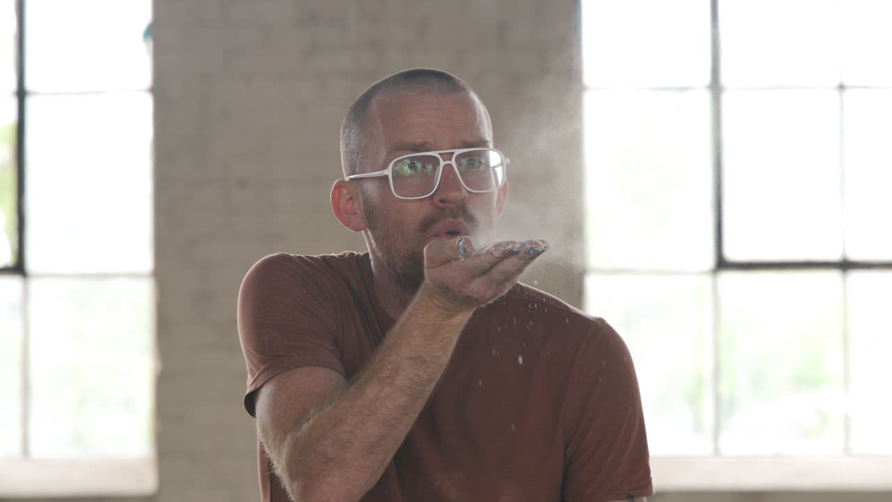 Man in an industrial warehouse setting blowing powder out of his hand.