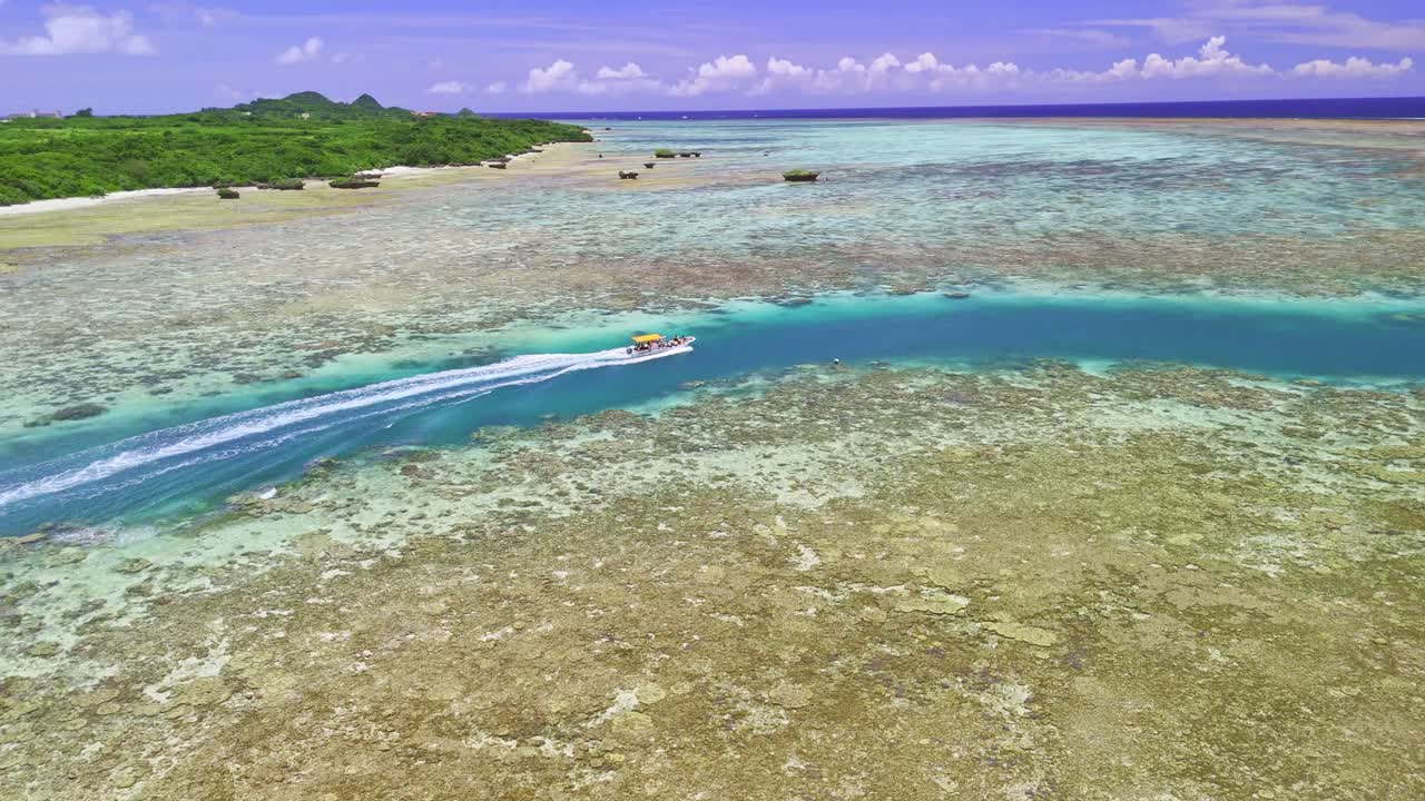 A stunning aerial shot captures a speedboat as it carves a path through a narrow, deep blue channel in the middle of a vast, shallow coral reef. The vibrant turquoise water contrasts beautifully