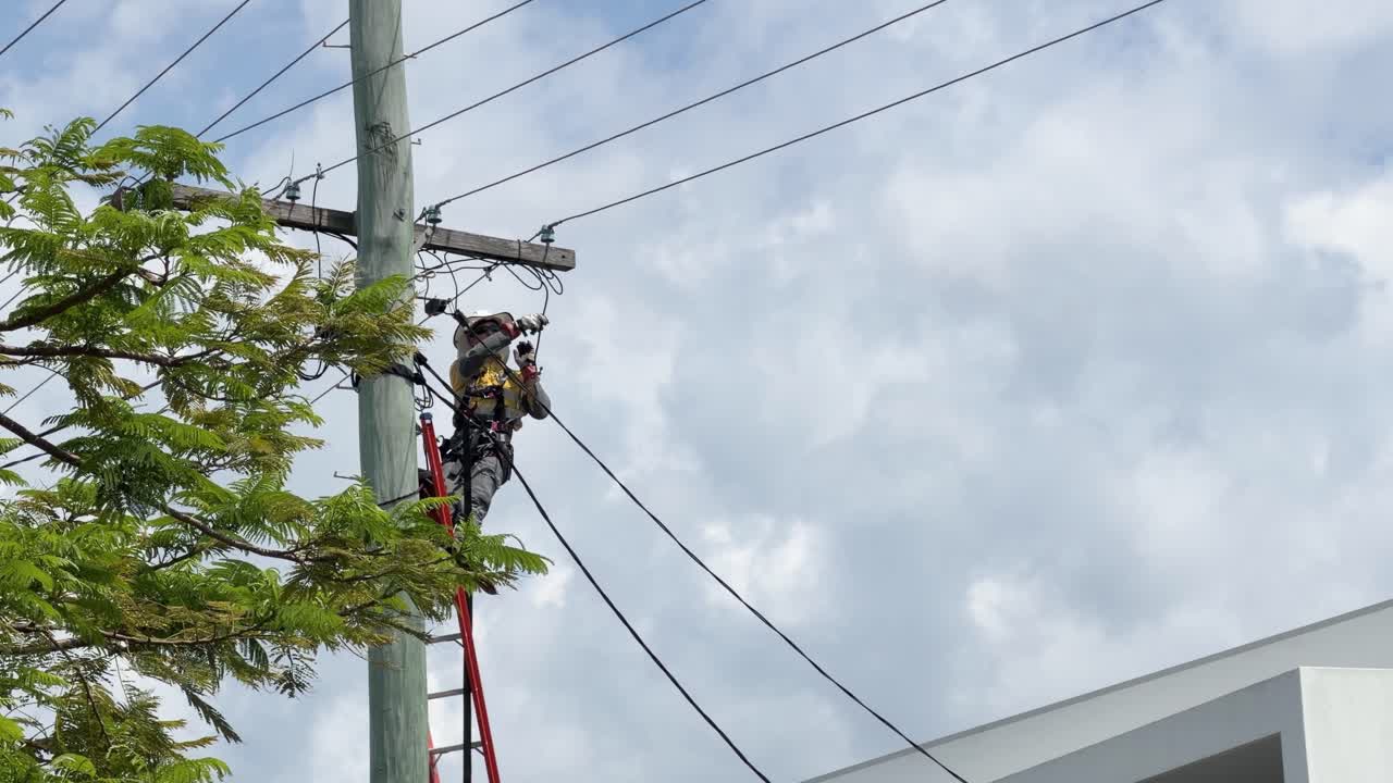 Utility worker repairs overhead power cables on ladder, wearing safety equipment, under bright daylight sky