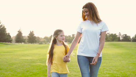 mother and little daughter holding hands and talking while walking on green grass field in the park