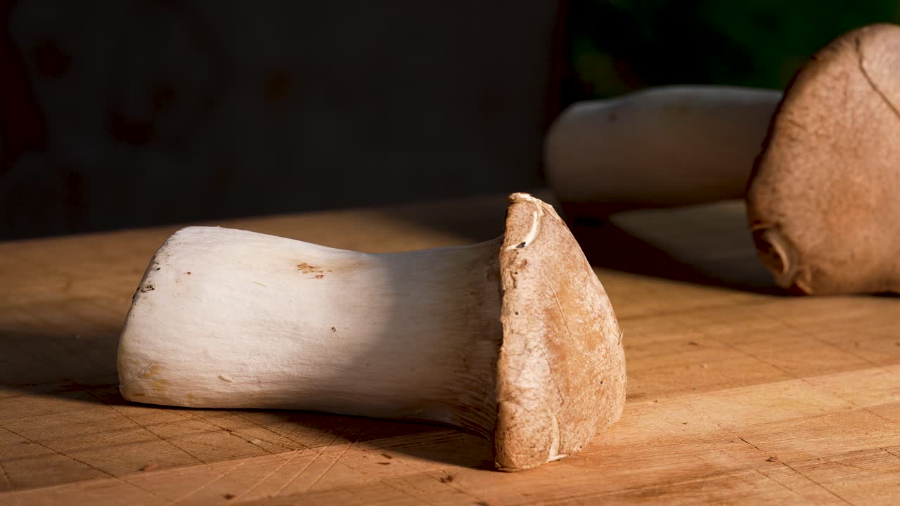 Close-up of a king oyster mushroom being cut cleanly in half with a sharp knife. The smooth motion reveals the dense texture and natural structure of the fresh mushroom