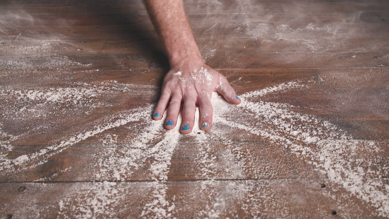 A close-up of a hand slamming down or brushing through a large cloud of white powder on a wooden surface, creating a dramatic burst or explosion of powder outward from the center.