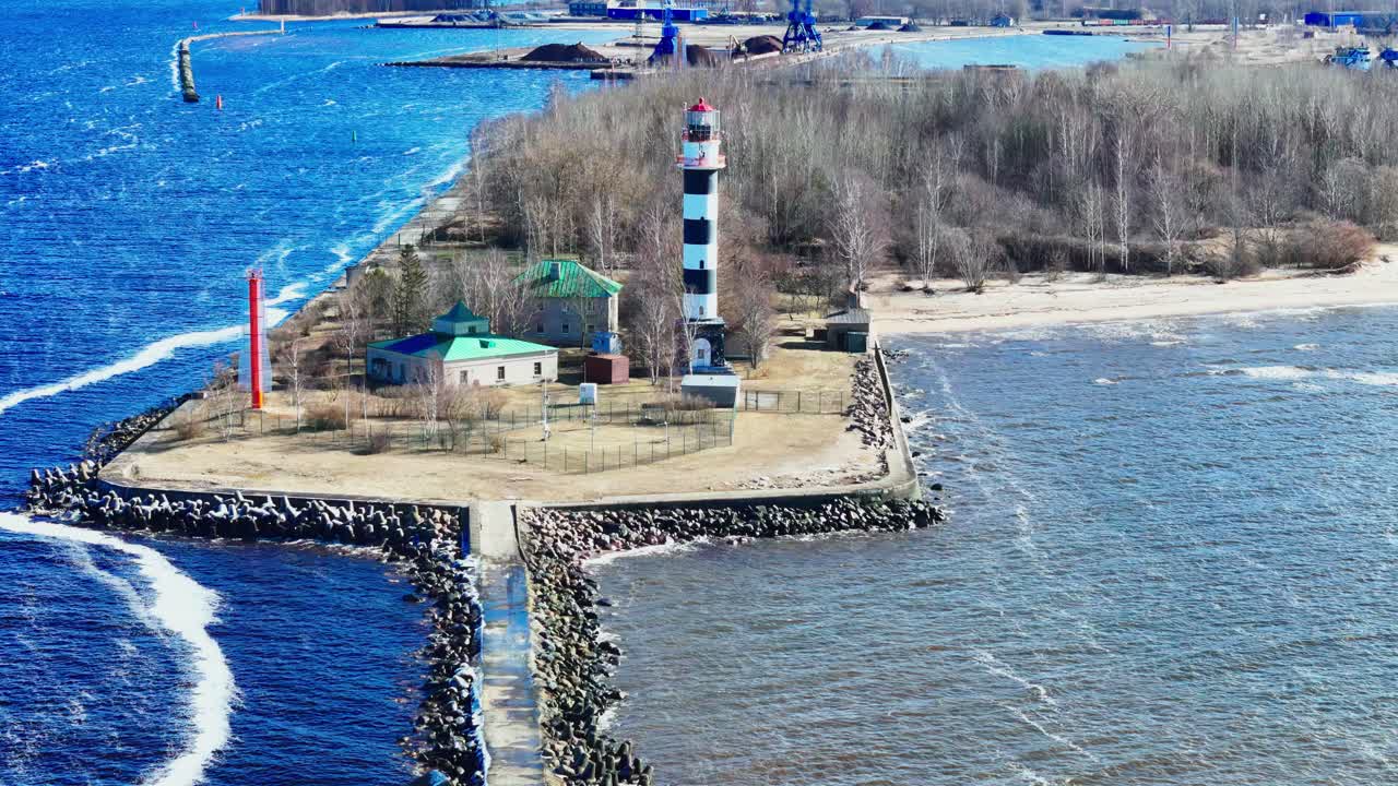 A black and white lighthouse rises above a slender stone jetty dividing bright blue sea from muted river waters, surrounded by rustic buildings, bare trees, and distant industry.