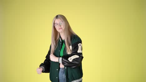 young woman in green varsity jacket and glasses posing against yellow background