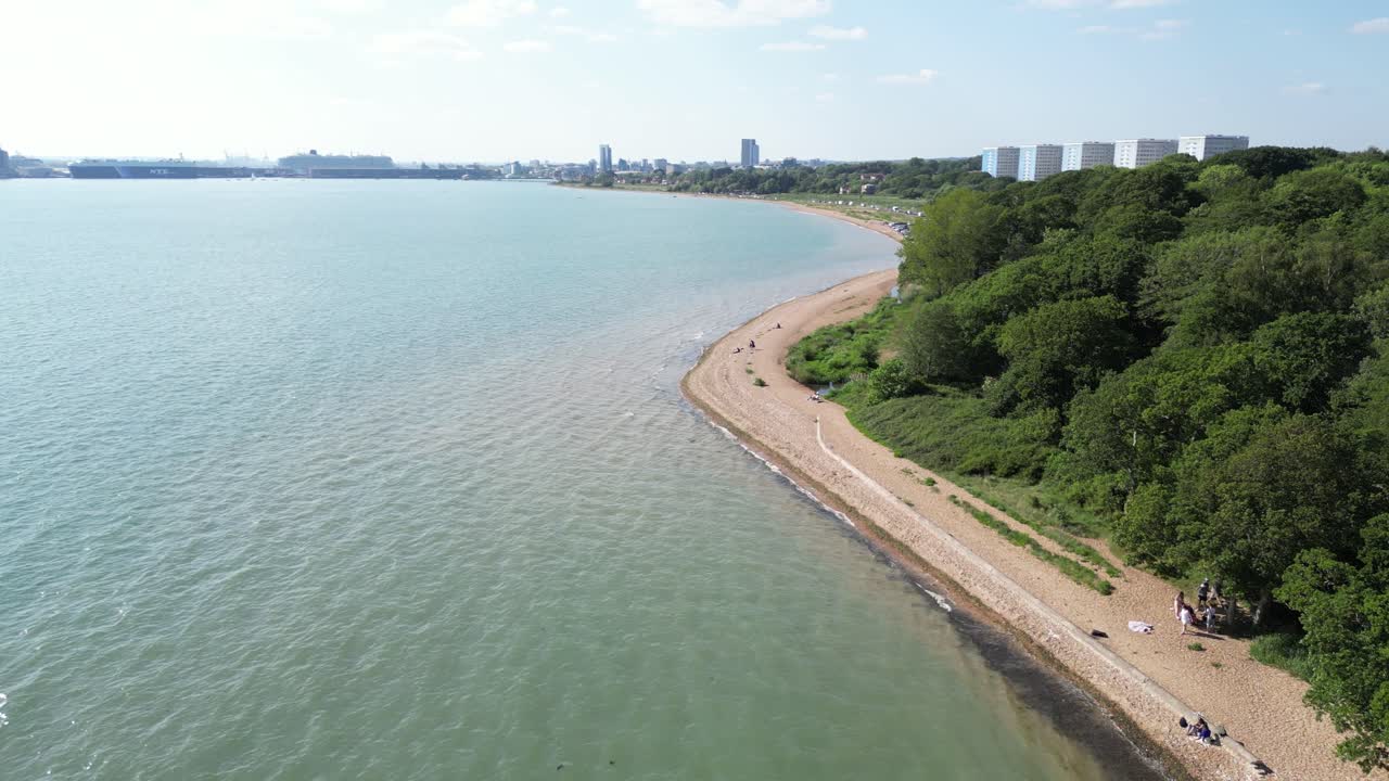 An aerial view captures the western shore of Southampton, featuring a sandy beach, lush greenery, and the city skyline in the distance under a clear sky.