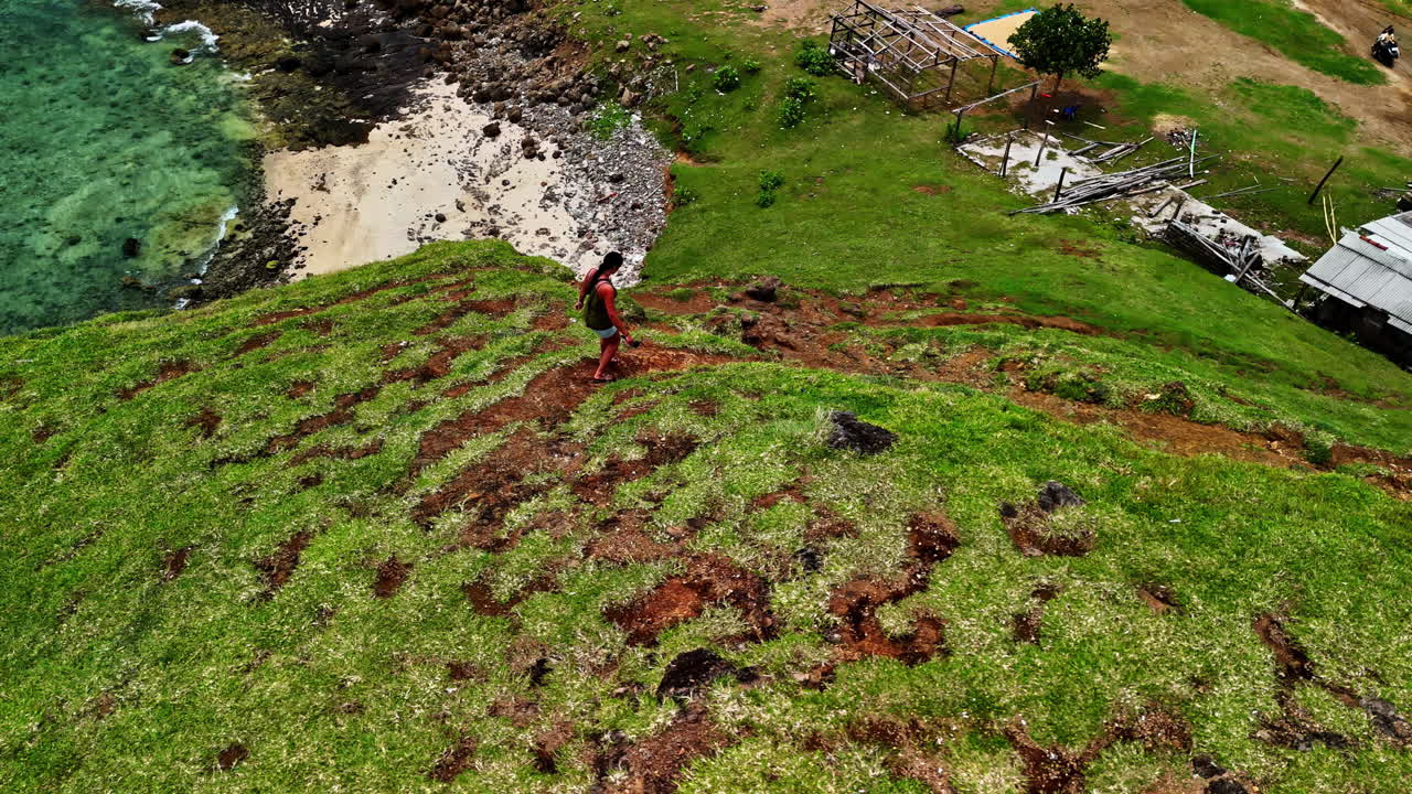From above Pantai Seger in Kuta Lombok, a woman stands on a grassy hill overlooking turquoise waters, white sands, and rocky shores with wooden structures nearby