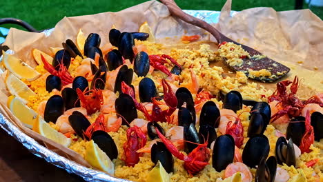 a close-up shot of a huge, freshly cooked pan of traditional spanish seafood paella with saffron rice, mussels, crayfish, and lemon wedges at a food festival