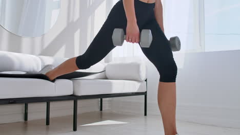 brunette in black sportswear in a white apartment makes a split squat with dumbbells in her hands leaning on the sofa with her foot.