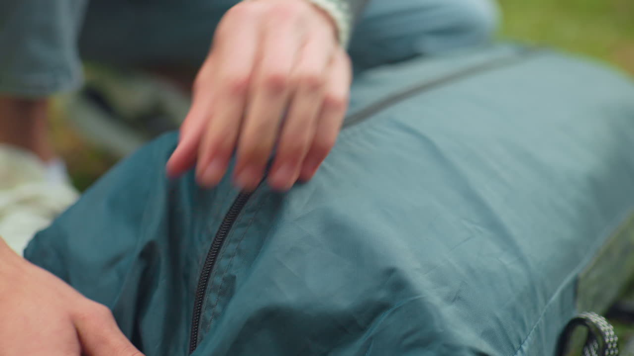 close up of person zipping camping bag filled with folded gear in forest environment, showing preparation for outdoor adventure with slight glimpse of hand