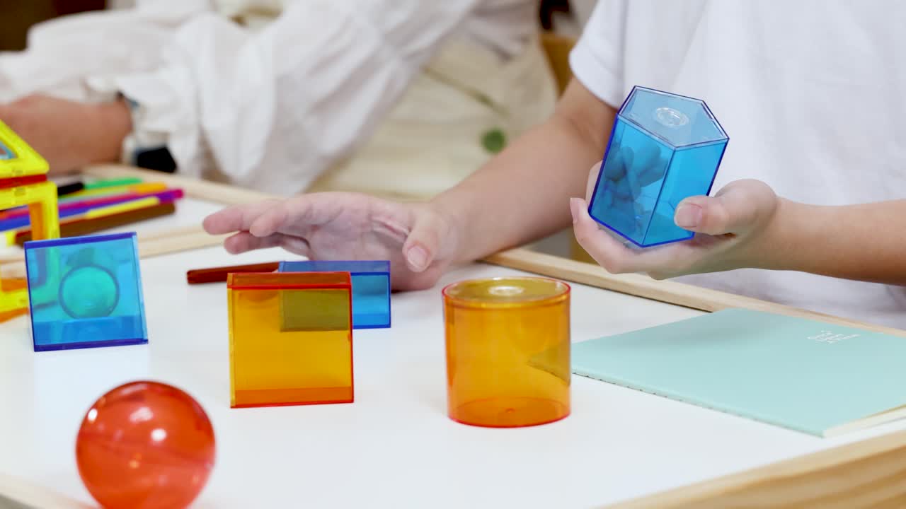 Student manipulates translucent geometric solids on desk in brightly lit classroom, emphasizing hands-on learning