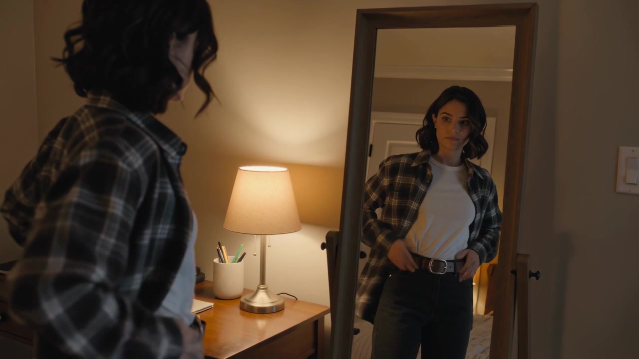 Una donna in camicia a quadri si sistema di fronte allo specchio, stringendo la cintura e completando l'outfit sotto la luce di una lampada da camera.