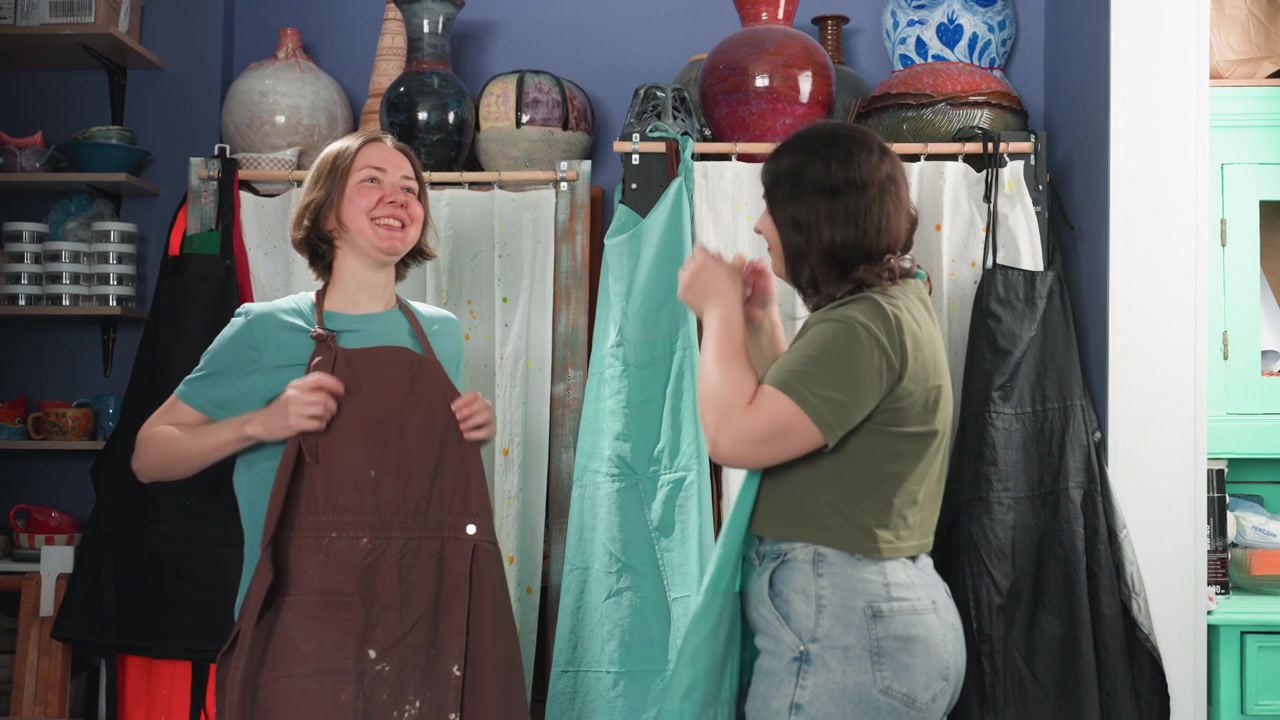 Prova di grembiuli in un laboratorio di ceramica: gioioso lavoro di squadra. Amici provano i grembiuli tra scaffali di vasi, uno sistema le cinghie mentre l'altro ride, condividendo eccitazione e un giocoso 'cinque'. Attrezzi da studio visibili, pronti