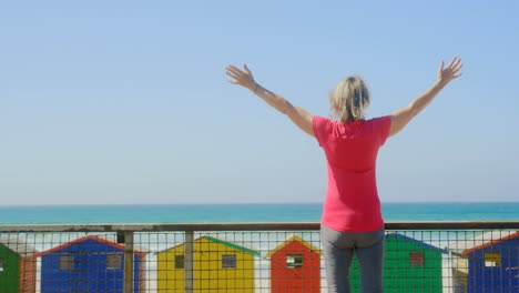 Rear-view-of-active-senior-Caucasian-woman-standing-with-arms-outstretched-on-promenade-at-beach-4k