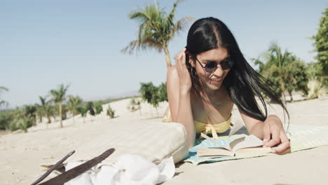 Feliz-Mujer-Hispana-Con-Gafas-De-Sol-Tumbada-En-La-Playa-Leyendo-Un-Libro-Al-Sol,-Copiando-Espacio,-Cámara-Lenta