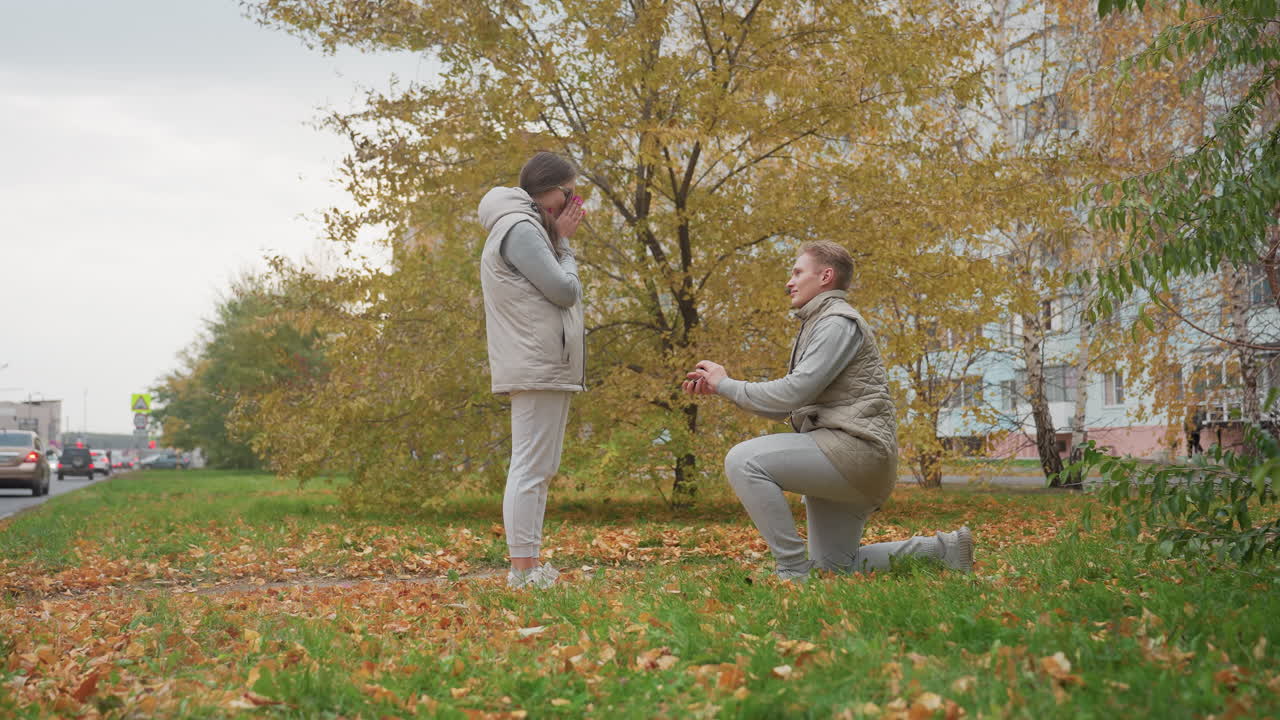 Young adult walks toward girlfriend preparing to kneel and propose in outdoor park setting, reacting with surprise as autumn leaves sway gently, background features trees, cars, and distant signpost