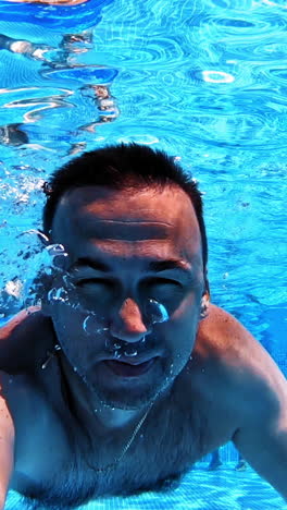 man swims underwater in the pool. blue clear water and a male swimming ahead with underwater camera. summer vacation at water. vertical video