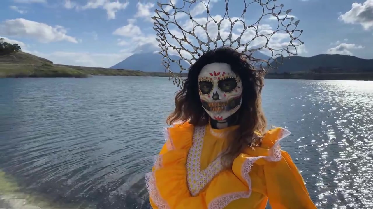 Telecamera puntata su una donna che posa come una Catrina messicana durante il Dia de Muertos, accanto a un lago in una giornata di sole.