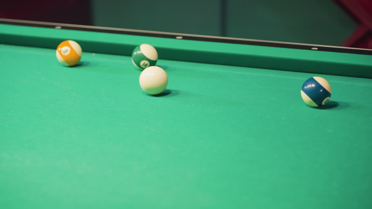 Close-up of person using cue stick to strike white billiard ball on green pool table. Focus on billiard balls scattered across table as shot is taken