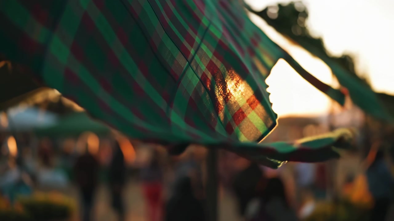 Tessuto scozzese in primo piano che attraversa diagonalmente la scena, con pieghe morbide, bordo sfrangiato, quadri verdi e rossi traslucidi, calda retroilluminazione del tramonto e forme di folla esterna fortemente sfocate sullo sfondo