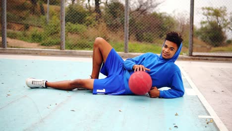 Young-african-american-lying-on-the-floor-of-the-playing-field-and-playing-with-basketball-ball.-Slow-Motion-shot