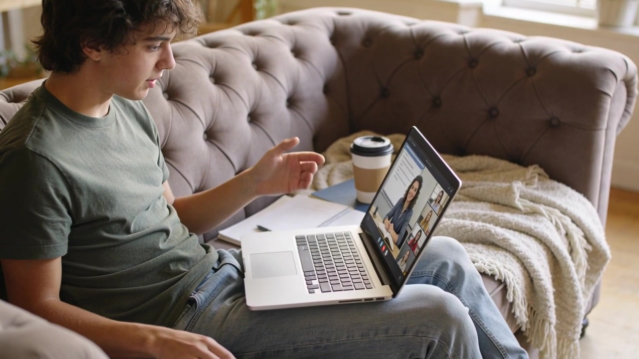Gesturing man in green tee jeans on tufted sofa responding to woman on laptop call, typing