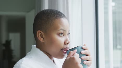 thoughtful african american plus size woman wearing robe, looking through window, drinking coffee