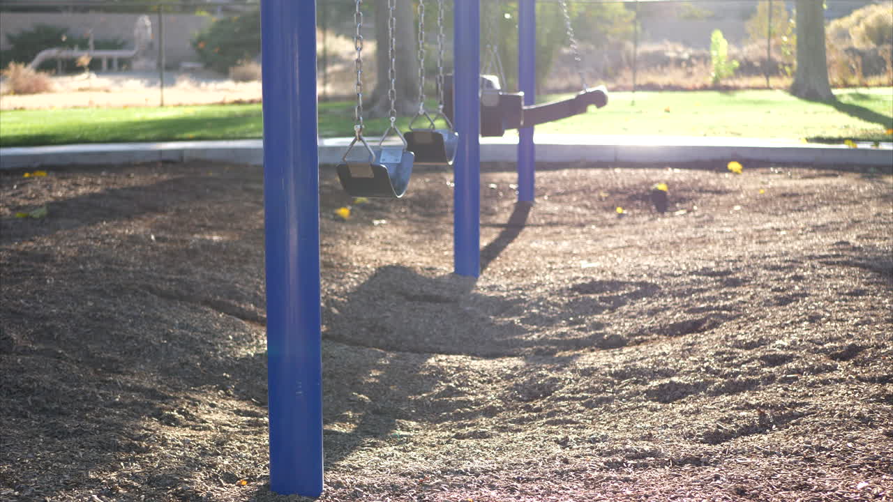 Slide left through a sunny kids park playground with an empty blue swing set