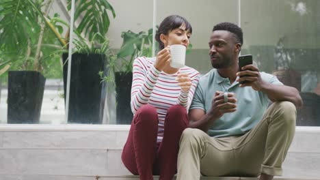 happy diverse couple wearing blouse and shirt talking and drinking coffee in garden