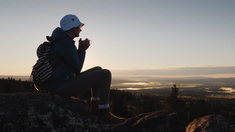 A-Traveler-Drinks-Tea-From-A-Mug-At-The-Peak-Of-The-Mountain-Sits-On-A-Rock-In-The-Background-A-Red-