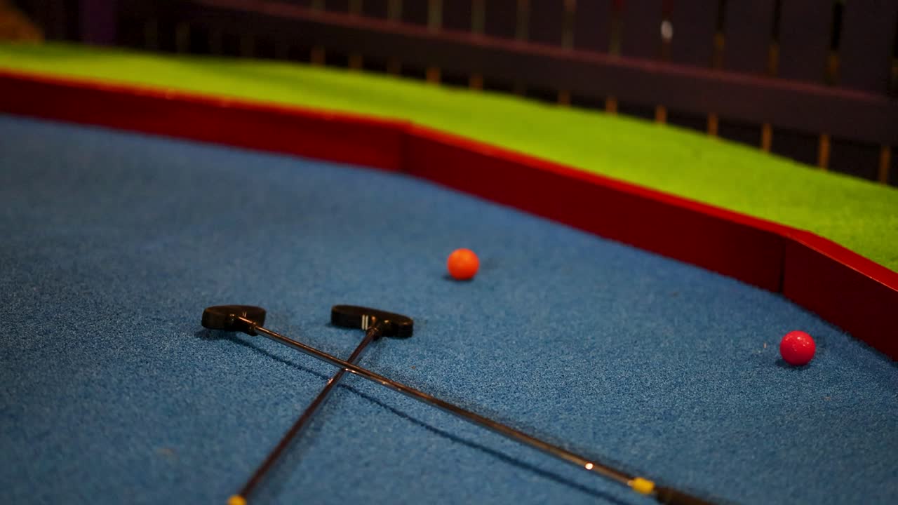 A golf ball bounces across a colorful indoor mini-golf course with two putters resting nearby
