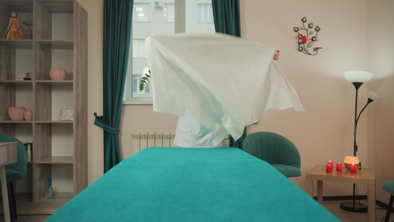 Massageterapeut klargør spa-behandlingsbriks til mikrostrømsbehandling, spreder sprødt hvidt lagen over petroleumsblåt tæppe, glatter hjørnerne, tænder stearinlys på sidebord, med vindueslys og hyggelig reol.