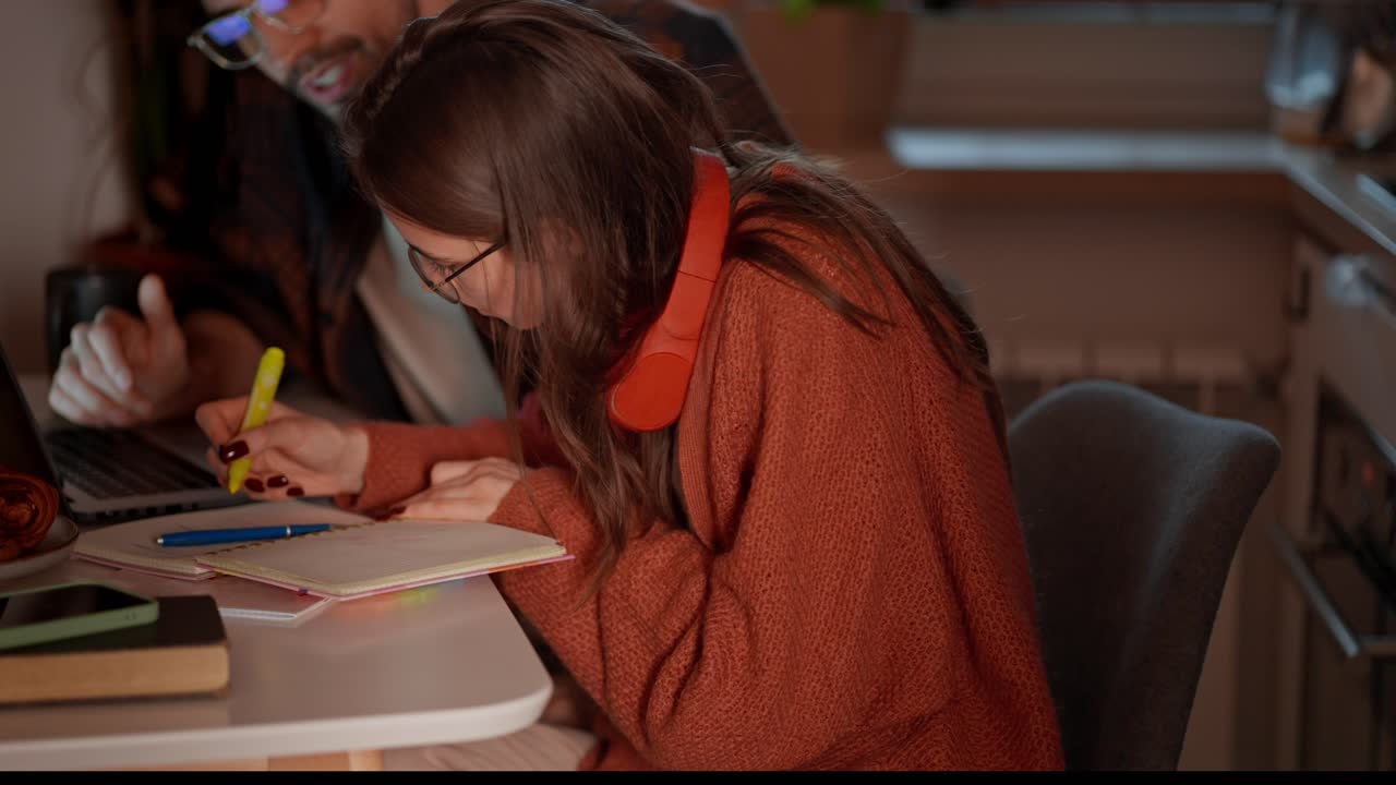 una joven morena segura con auriculares rojos inalámbricos y un suéter naranja estudia lenguas extranjeras con su maestra morena y toma notas en sus notas mientras estudia idiomas extranjeros en un apartamento moderno en la cocina