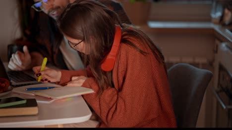 a young confident brunette girl in red wireless headphones and an orange sweater studies foreign languages with her teacher brunet and takes notes in her notes while studying foreign languages in a modern apartment in the kitchen