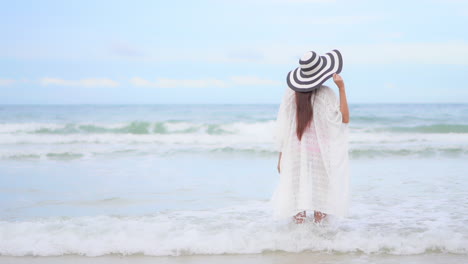 은폐를 입은 젊은 여성이 서핑이 그녀의 다리에 부딪칠 때 카메라에 등을 대고 서 있습니다.