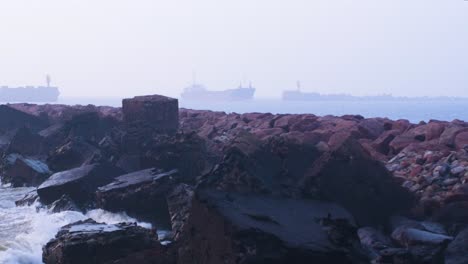 large blue cargo ship entering the port of liepaja in foggy day, stone pier in foreground, waves splashing, distant medium shot