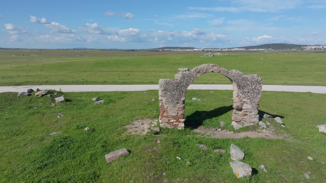 Lateral drone flight over green pastures featuring the old “Puerta de Arenas,” a brick and stone archway, remains of a rural structure with historical value in open countryside.